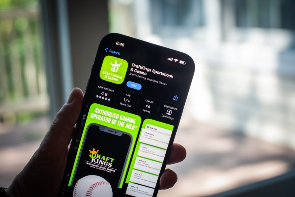Mão segurando smartphone com o aplicativo DraftKings Sportsbook & Casino em destaque na loja de apps.