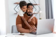 um pai usando o notebook enquanto o filho o abraça pelas costas