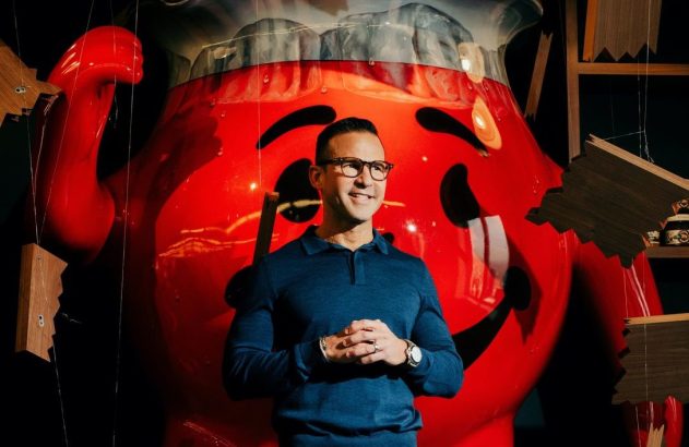 Homem sorrindo em pé, com as mãos juntas, na frente do mascote gigante Kool-Aid Man.
