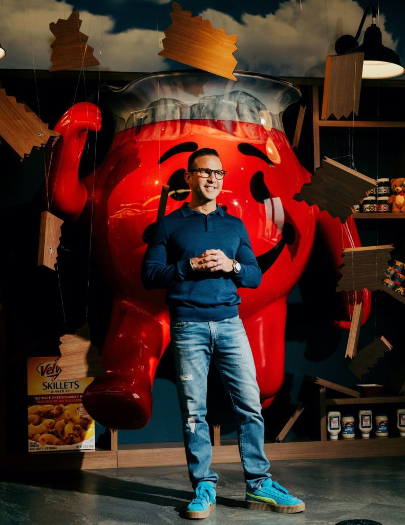 Homem sorridente à frente de jarro gigante do Kool-Aid Man. Caixa 'Vely Skillets' e garrafas de maionese visíveis.
