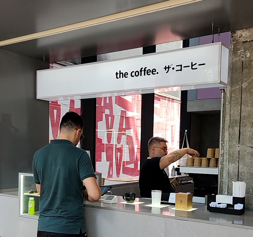 Dois homens em balcão de café: um cliente pede, o barista serve. Letreiro "the coffee" acima.