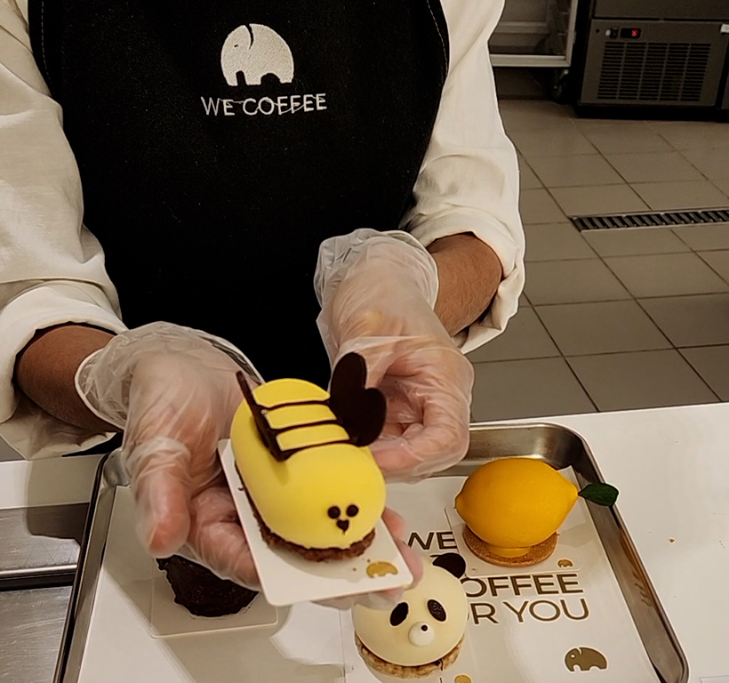 Pessoa de avental WE COFFEE e luvas apresenta um doce amarelo em forma de abelha. Bandeja com doces.