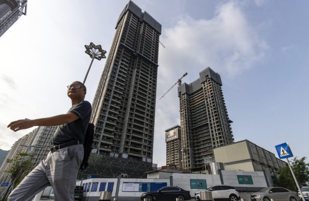 Homem andando em rua urbana. Prédios altos em construção dominam o fundo. Céu claro e carros na base.
