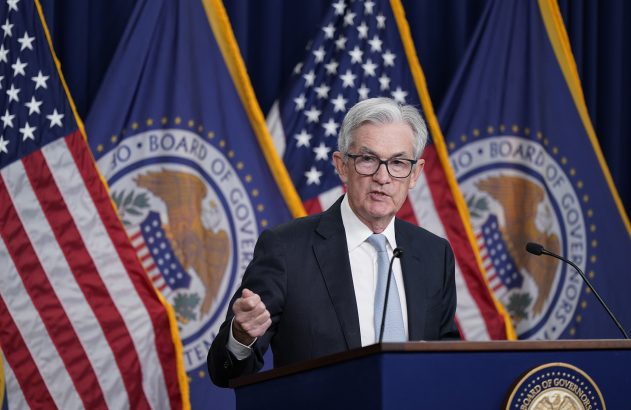 Homem de terno fala em pódio com bandeiras americanas e do Federal Reserve ao fundo.