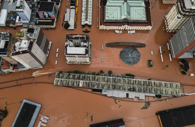 Vista aérea de cidade com ruas e edifícios inundados por água marrom, mostrando os estragos de uma enchente.