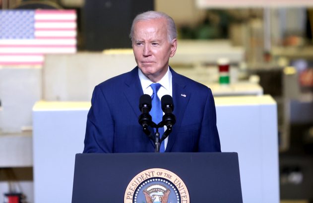 Joe Biden em terno azul discursa em pódio com selo presidencial; bandeira dos EUA ao fundo.