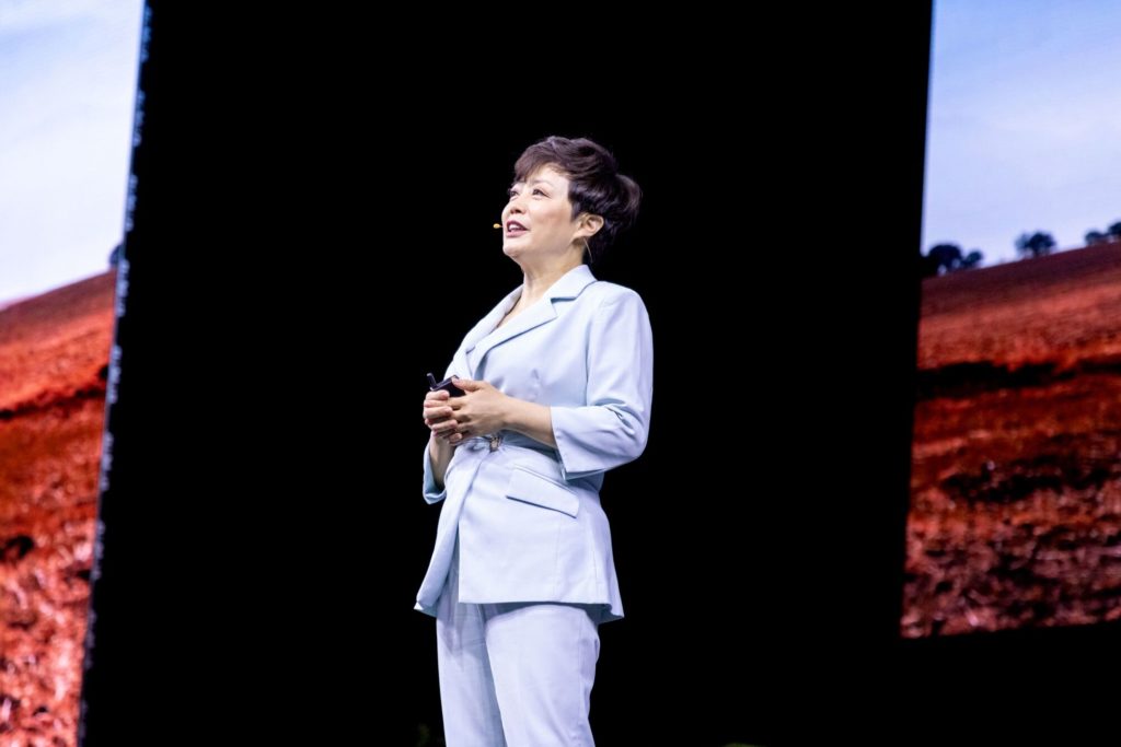 Mulher em terno azul claro apresenta em palco. Telas laterais exibem paisagens de campo com céu.
