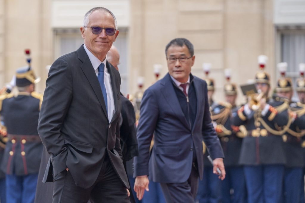 Dois homens de terno andam. Um sorri, de óculos. Ao fundo, guardas em uniforme formal.