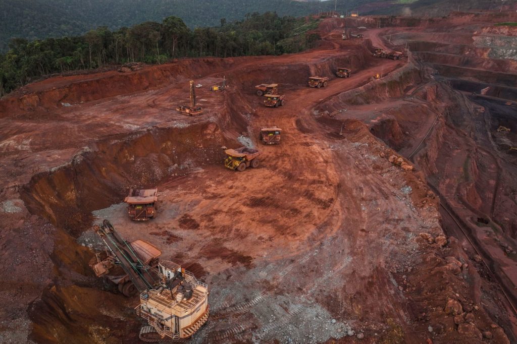 Mina a céu aberto com escavadeira e vários caminhões transportando minério. Terra vermelha e floresta ao fundo.