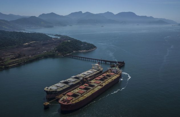 Navios graneleiros em porto industrial, com pátio de minério, montanhas e mar.