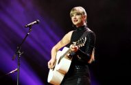 Taylor Swift tocando violão branco em palco com vestido preto cintilante e microfone.
