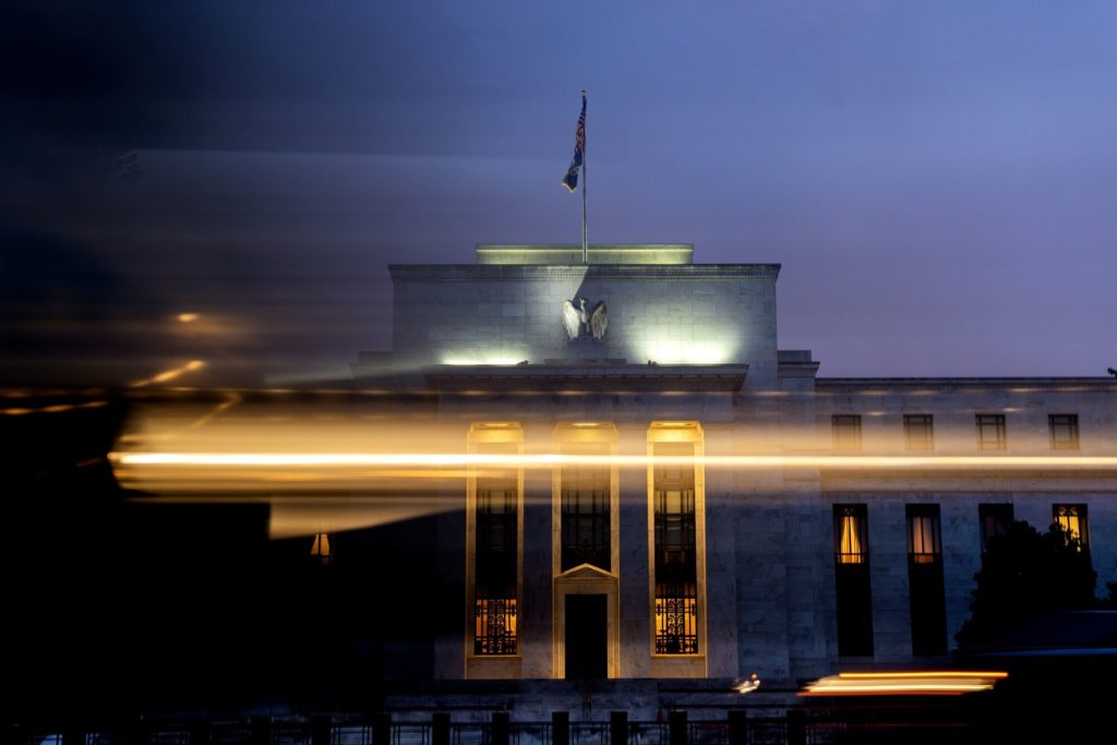 Edifício Federal Reserve à noite, com fachada iluminada, bandeira, águia e rastros de luz de veículos.