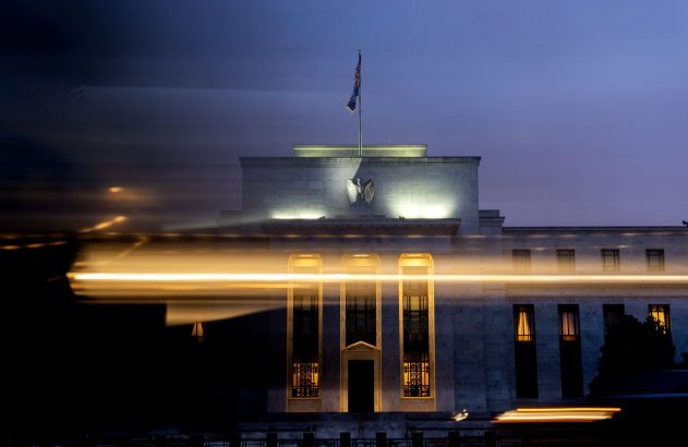 Edifício Federal Reserve à noite, com fachada iluminada, bandeira, águia e rastros de luz de veículos.