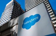 Sinal grande branco com o logo azul da Salesforce e edifícios modernos ao fundo.
