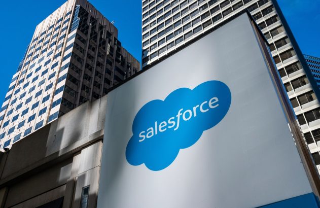 Sinal grande branco com o logo azul da Salesforce e edifícios modernos ao fundo.