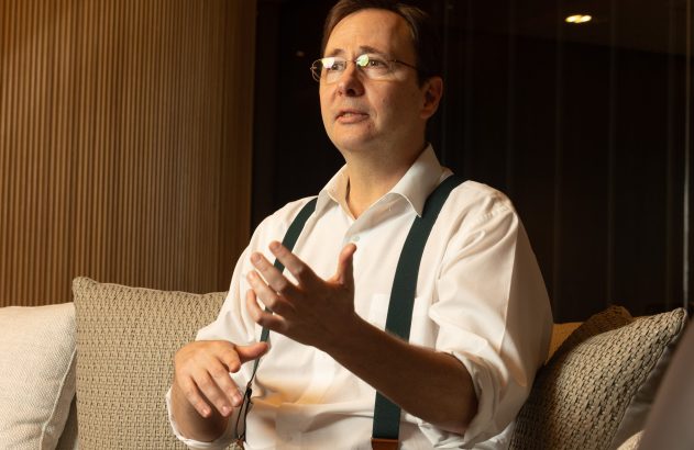 Homem de óculos, camisa branca e suspensórios, sentado em sofá, gesticula com as mãos enquanto conversa.