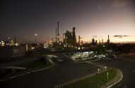 Vista noturna de refinaria industrial com estruturas iluminadas e céu avermelhado. Estrada em primeiro plano.