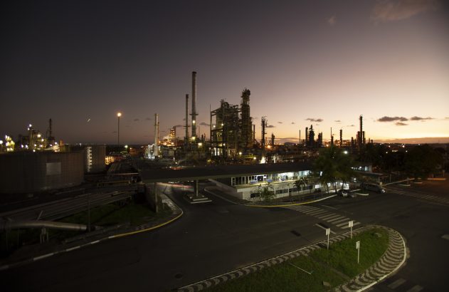 Vista noturna de refinaria industrial com estruturas iluminadas e céu avermelhado. Estrada em primeiro plano.