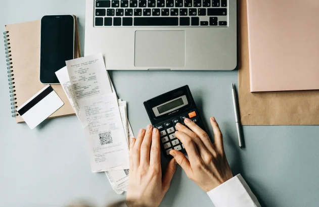 Mão de mulher calculando, usando calculadora, em mesa com notebook, anotações e notas fiscais