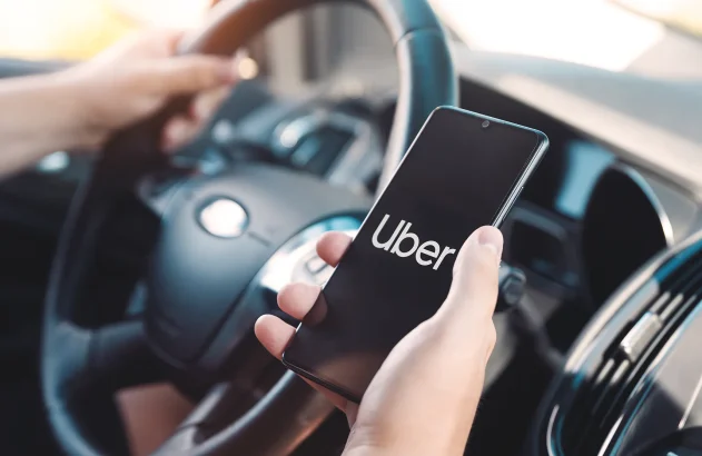 IMagem de um motorist segurando o volante com a mão esquerda e com um celular mostrando o logo da Uber na direita