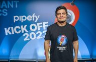 Homem sorridente em camiseta ‘Brilliant Basics’ da Pipefy, em frente ao painel ‘KICK OFF 2023’.