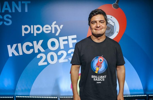 Homem sorridente em camiseta ‘Brilliant Basics’ da Pipefy, em frente ao painel ‘KICK OFF 2023’.