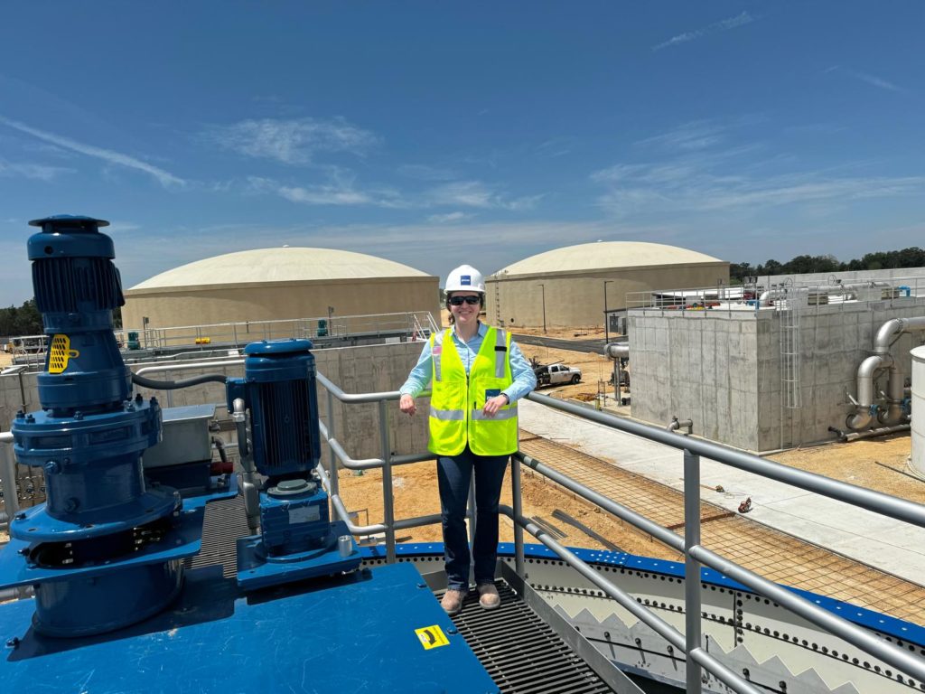 Mulher com capacete e colete de segurança em plataforma elevada de planta industrial sob céu azul.