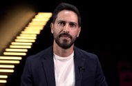 Homem barbudo de blazer escuro e camiseta branca, olhando à frente, com microfone de lapela. Fundo preto com listras luminosas.