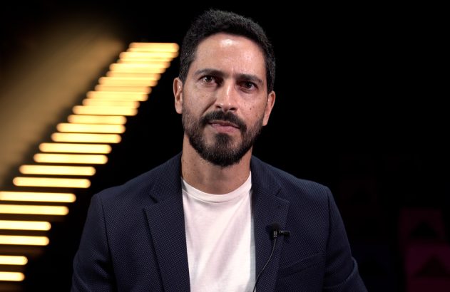 Homem barbudo de blazer escuro e camiseta branca, olhando à frente, com microfone de lapela. Fundo preto com listras luminosas.
