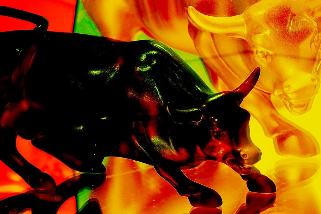 Duas esculturas de touros, um escuro e um dourado, em pose de ataque sobre superfície reflexiva e fundo vibrante.