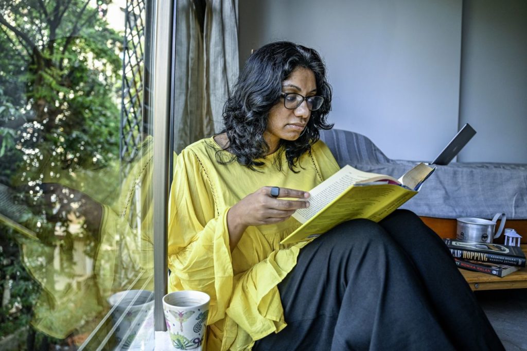 Mulher de óculos sentada no chão, lendo um livro amarelo junto a uma janela.