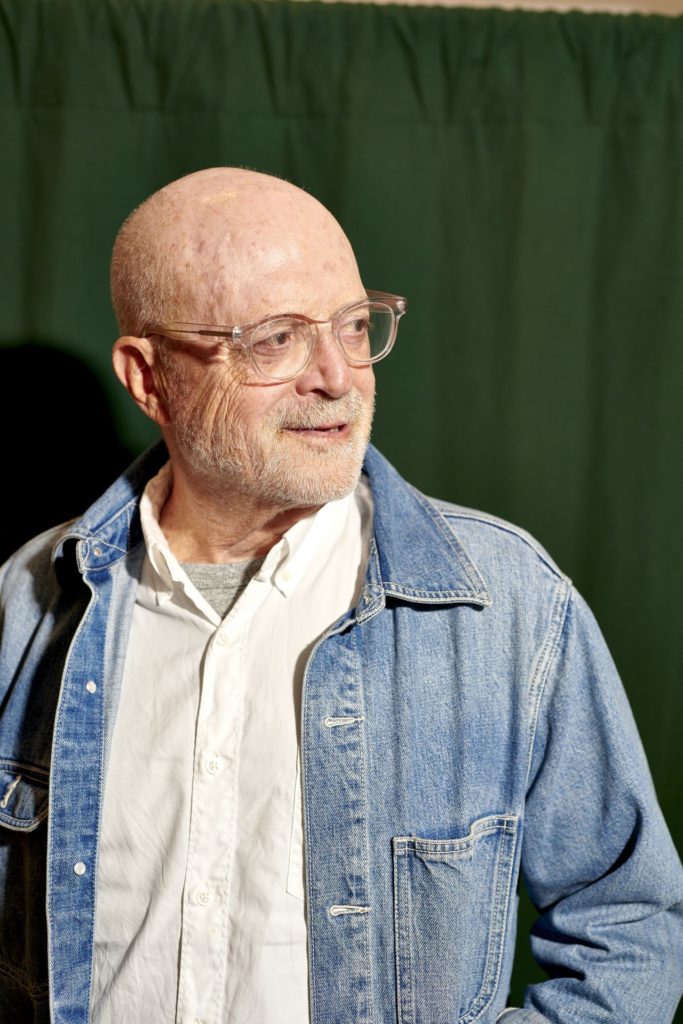 Retrato de um homem careca com barba e óculos, sorrindo e olhando para o lado. Ele veste jaqueta jeans e camisa branca, contra um fundo verde.