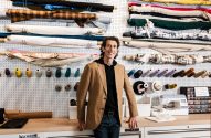 Homem sorridente em estúdio de costura, cercado por rolos de tecido, carretéis de linha e máquina de costura.