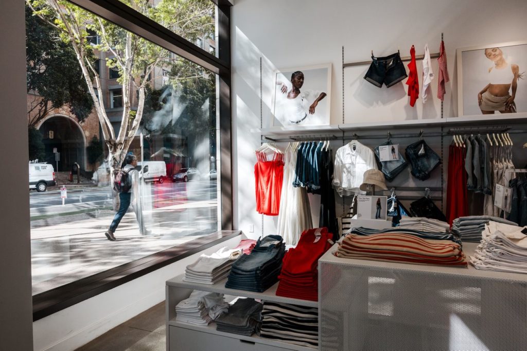 Loja de roupas com vitrine ampla, exibindo peças de vestuário. Pedestre refletido na janela da rua.