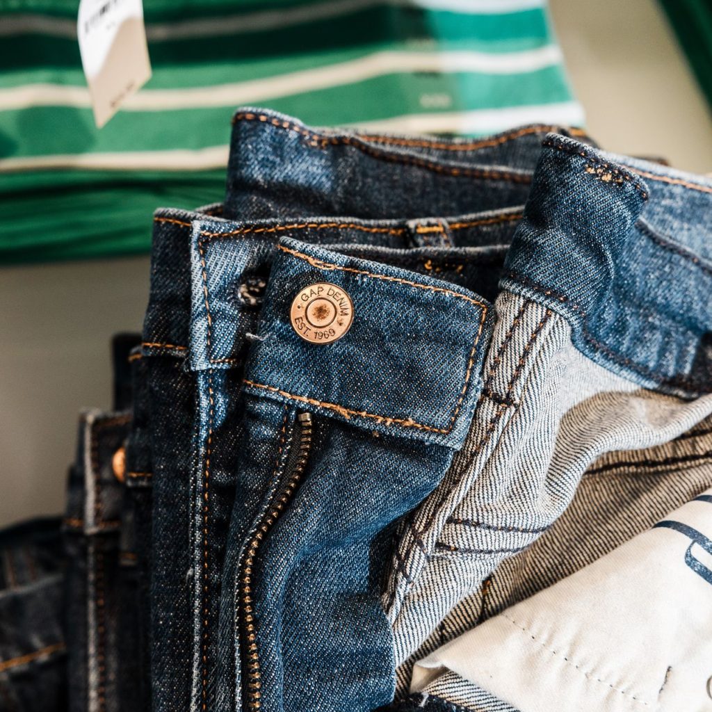 Jeans azuis dobrados com botão "GAP DENIM EST. 1969". Fundo desfocado com camiseta verde listrada.