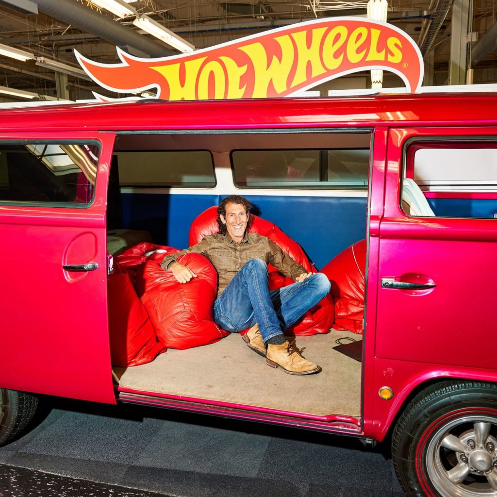 Homem sorridente sentado em pufes vermelhos em uma Kombi vermelha com logo Hot Wheels no teto.