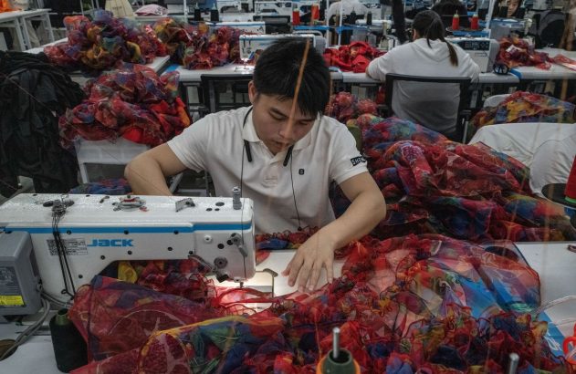 Em fábrica, homem costura tecido multicolorido vibrante em máquina industrial Jack, cercado por pilhas de material.