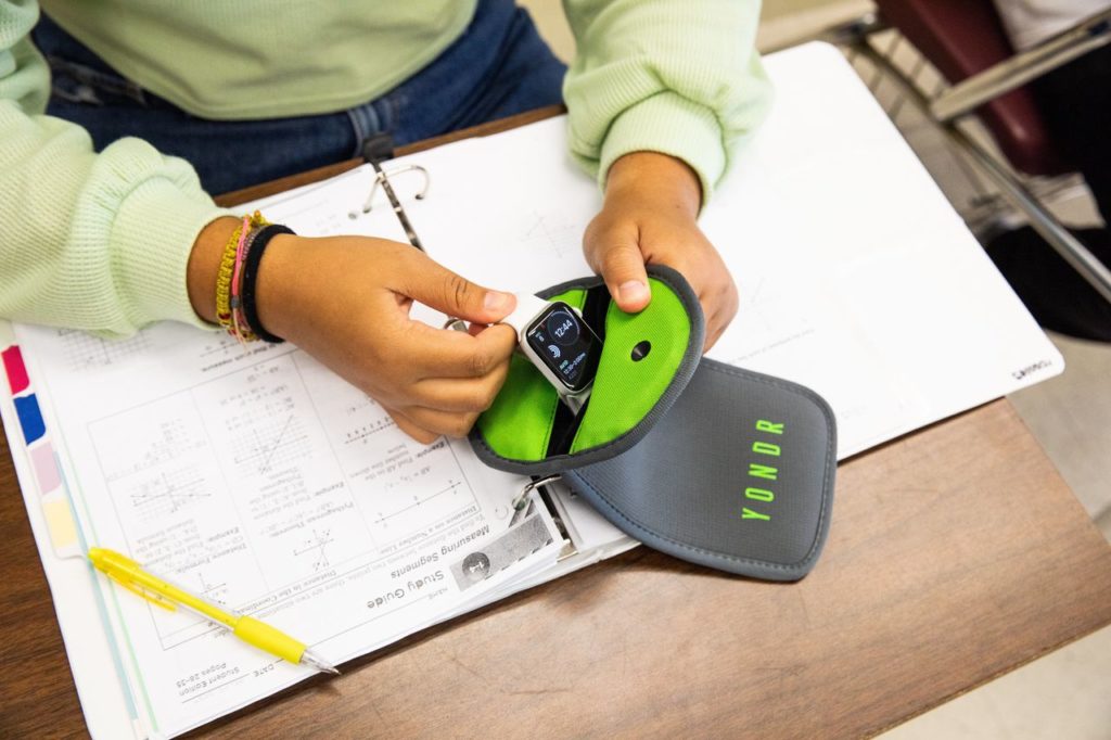 Mãos de aluno guardando smartwatch em bolsa Yondr sobre a carteira.