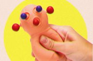 Mão apertando um brinquedo sensorial rosa claro com esferas vermelhas e azuis. Fundo amarelo.