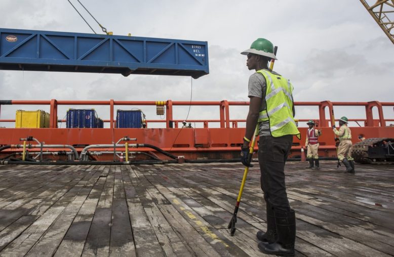Operário com colete em convés de navio observa guindaste içar contêiner azul.