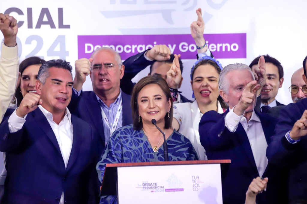 Mulher fala em pódio; pessoas atrás levantam punhos e cruzam dedos, em debate presidencial.