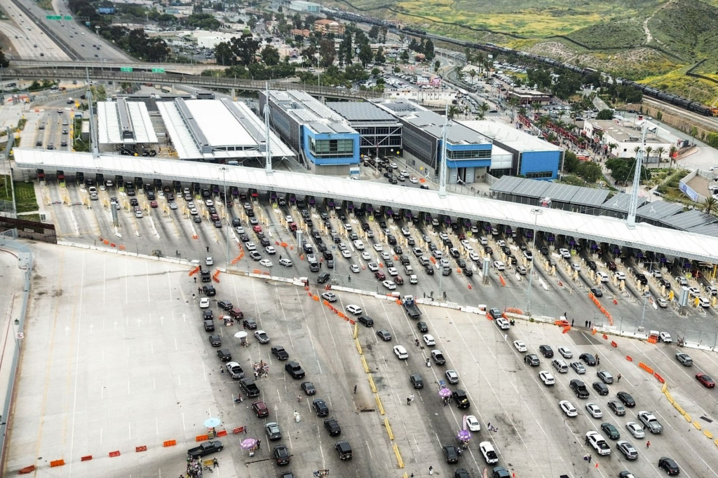 Vista aérea de posto de fronteira com inúmeras filas de veículos aguardando inspeção. Cenário urbano e natural.