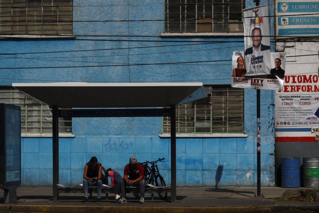 Pessoas sentadas em ponto de ônibus 'POPOCATEPETL' em frente a prédio azul, com cartazes políticos.