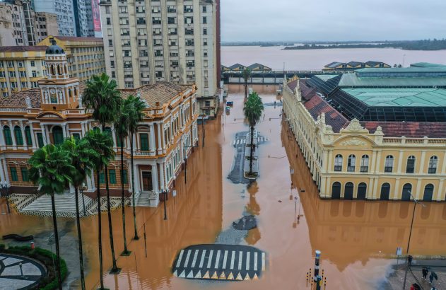 Rua de cidade histórica com prédios e palmeiras, completamente inundada por água barrenta e marrom.