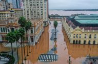 Cidade inundada: ruas e parte de edifícios submersos em água barrenta. Palmeiras e rio ao fundo sob céu nublado.