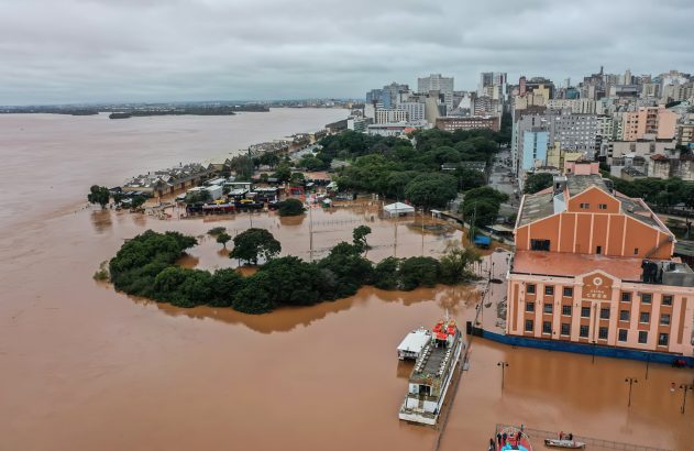 Vista aérea de cidade sob forte inundação. Água barrenta cobre ruas e prédios, barcos flutuam. Céu nublado.
