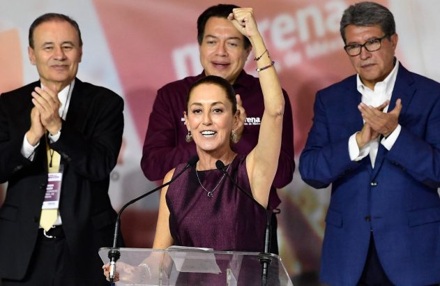 Mulher no pódio com punho erguido, dois homens batem palmas atrás dela em evento político.