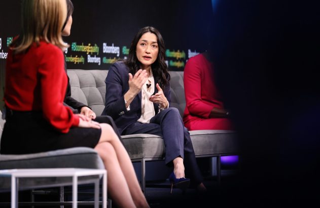 Mulher em painel da Bloomberg gesticula ao falar, outras participantes sentadas em um sofá no palco.