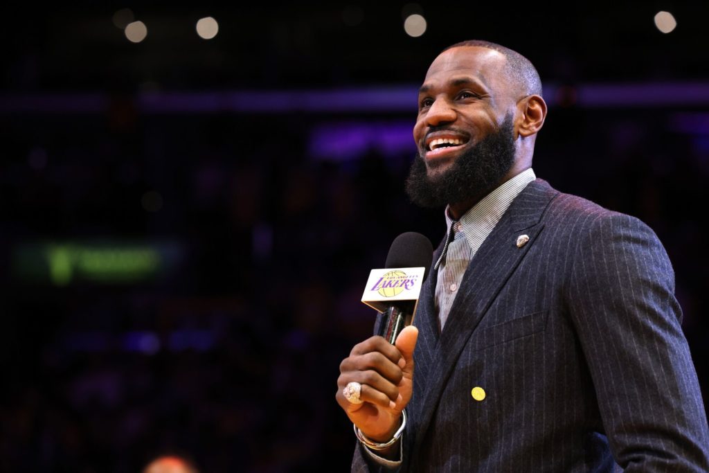 LeBron James, sorrindo, fala ao microfone com logo dos Lakers em uma arena escura.