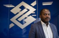 Homem negro em terno azul, olhando para câmera. Ao fundo, logo Banco do Brasil em branco sobre parede azul.
